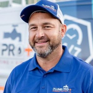 John Greider, Owner, wearing a blue Plumb Pro shirt and cap smiles proudly in front of a blurred company vehicle, ready to serve Goose Creek residents with top-notch plumbing and HVAC services.